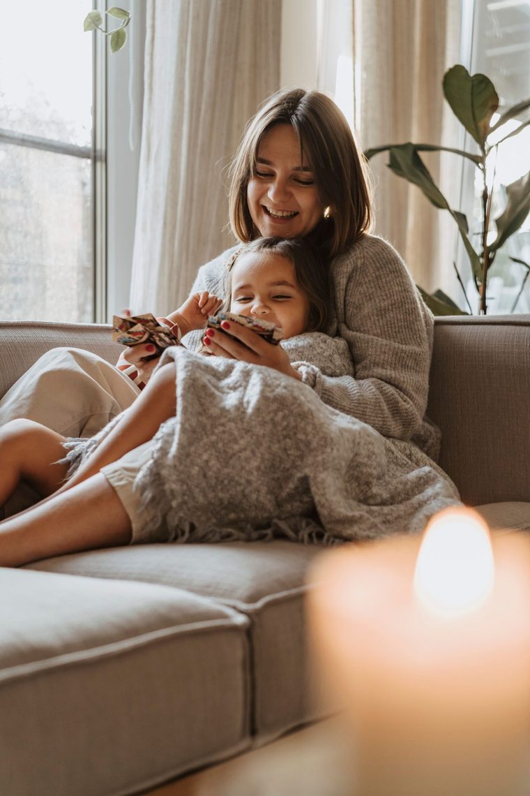 A smiling mother and daughter under a blanket on the sofa in a bright living room, looking at a colourful picture book together, with a candle burning in front of them