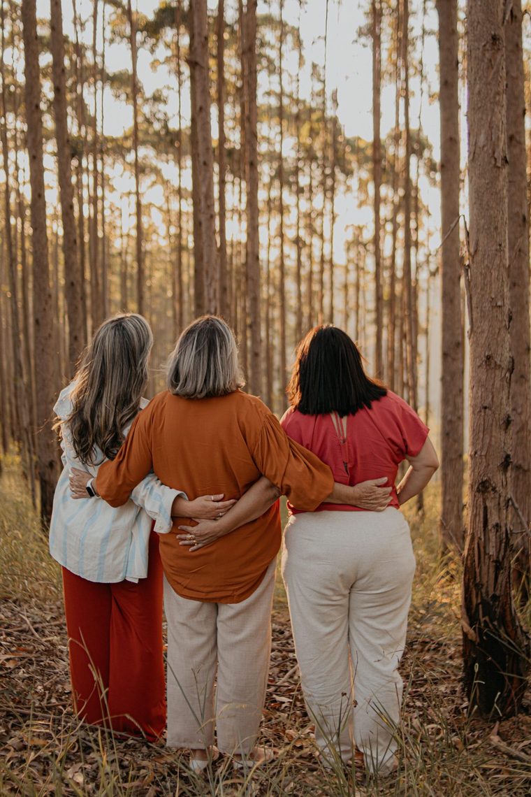 Three women seen from behind, arms around each other's waists, standing together in a pine forest in the warm light of the setting sun